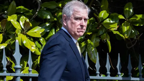 Getty Images Andrew Mountbatten-Windsor walking outside in a suit looking at the camera on a sunny day. Railings and hedging behind him