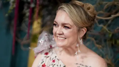 Colleen Hoover at a film premiere, wearing a jeweled white one-shoulder dress with her hair in an up-do and white feather earrings.