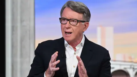 BBC Close-up of Peter Mandelson, wearing a black jacket and white button-up shirt and black spectacles, speaking at a BBC studio.