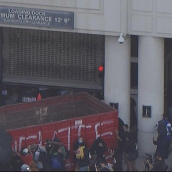 A crowd pushes a large construction dumpster and blocks the federal building's loading dock. - cbs