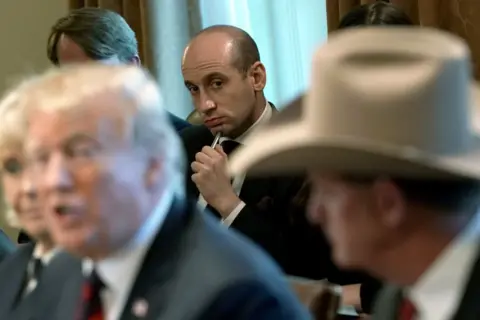 Getty Images Stephen Miller is in the background but sharply in focus. He is holding a pen and looking at Trump who is speaking in the foreground.
