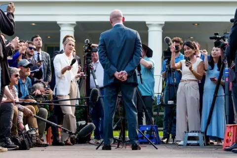 Getty Images Miller's back is to the camera and he is wearing a blue suit, stood facing reporters and microphones and cameras in front of the White House