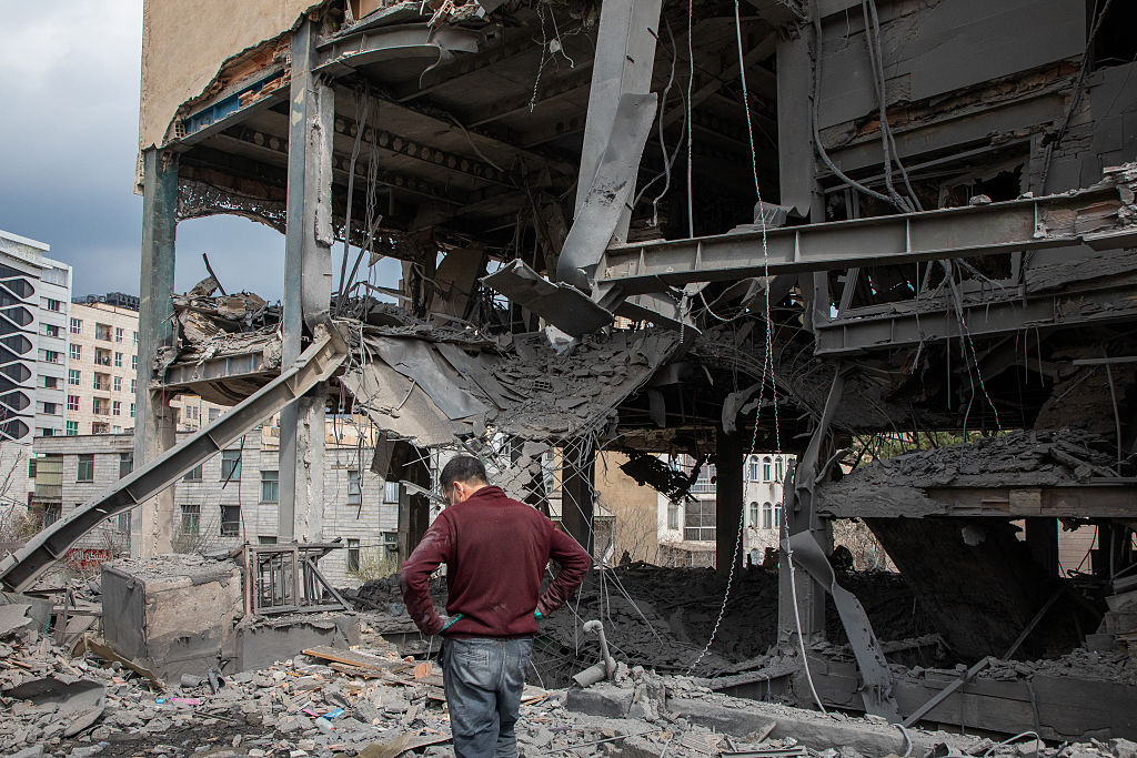 War-damaged buildings with debris in the street as a lone person walks through the scene.